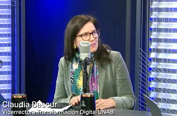 Claudia Basur, Vice Rector of Digital Information at UNAB, sits at a studio desk with a microphone, speaking on air with mugs nearby.