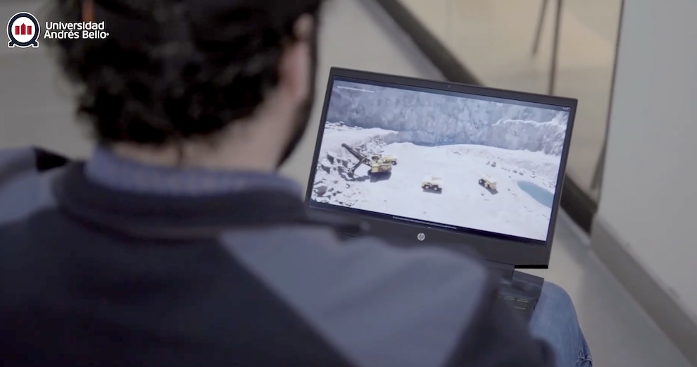 Back view of a person wearing a dark jacket, watching a laptop displaying a snowy quarry scene with trucks; Universidad Andrés Bello logo in corner.